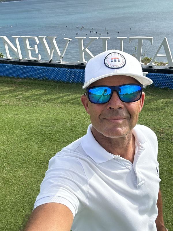 Selfie av Bobby med hvit caps og solbriller foran New Kuta-skiltet ved havet